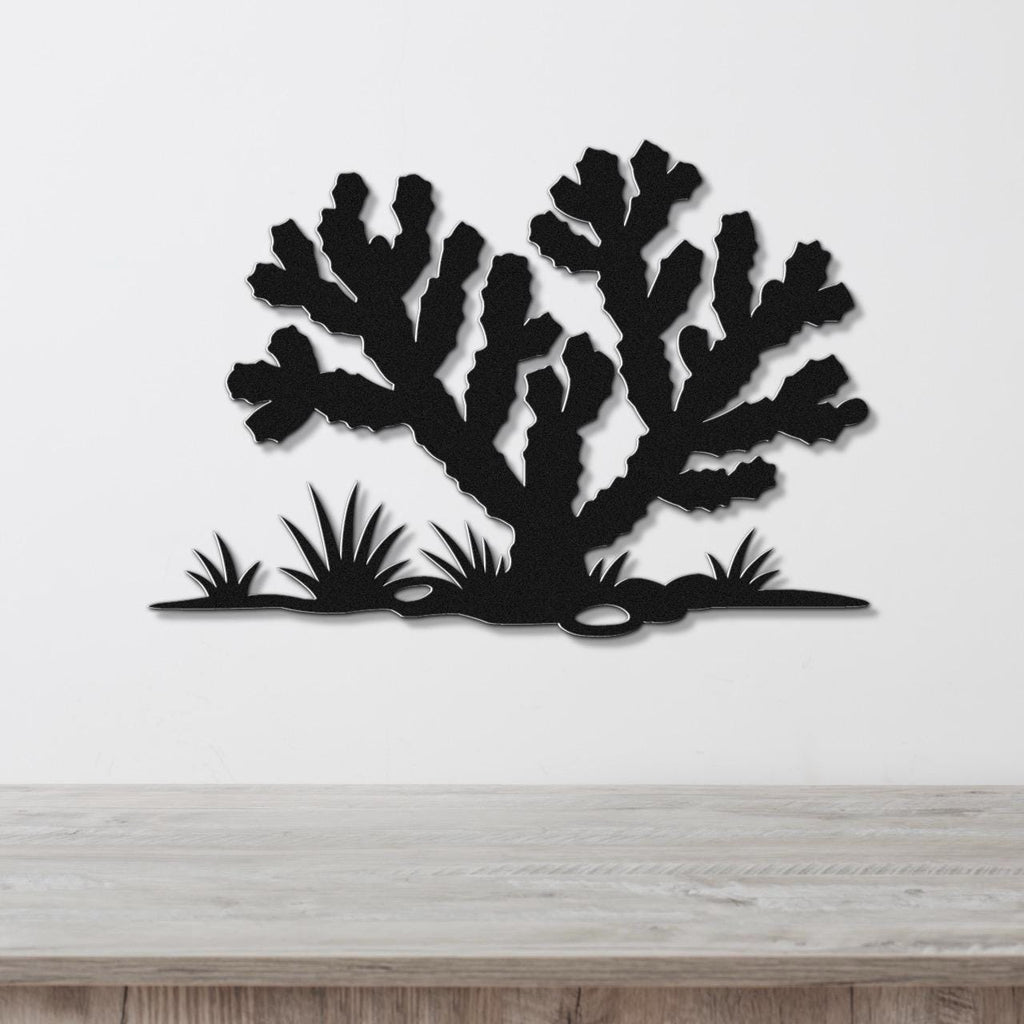 Black metal wall art cutout of a cholla cactus with spiky branches and desert plants, mounted on a white wall above a wooden surface.