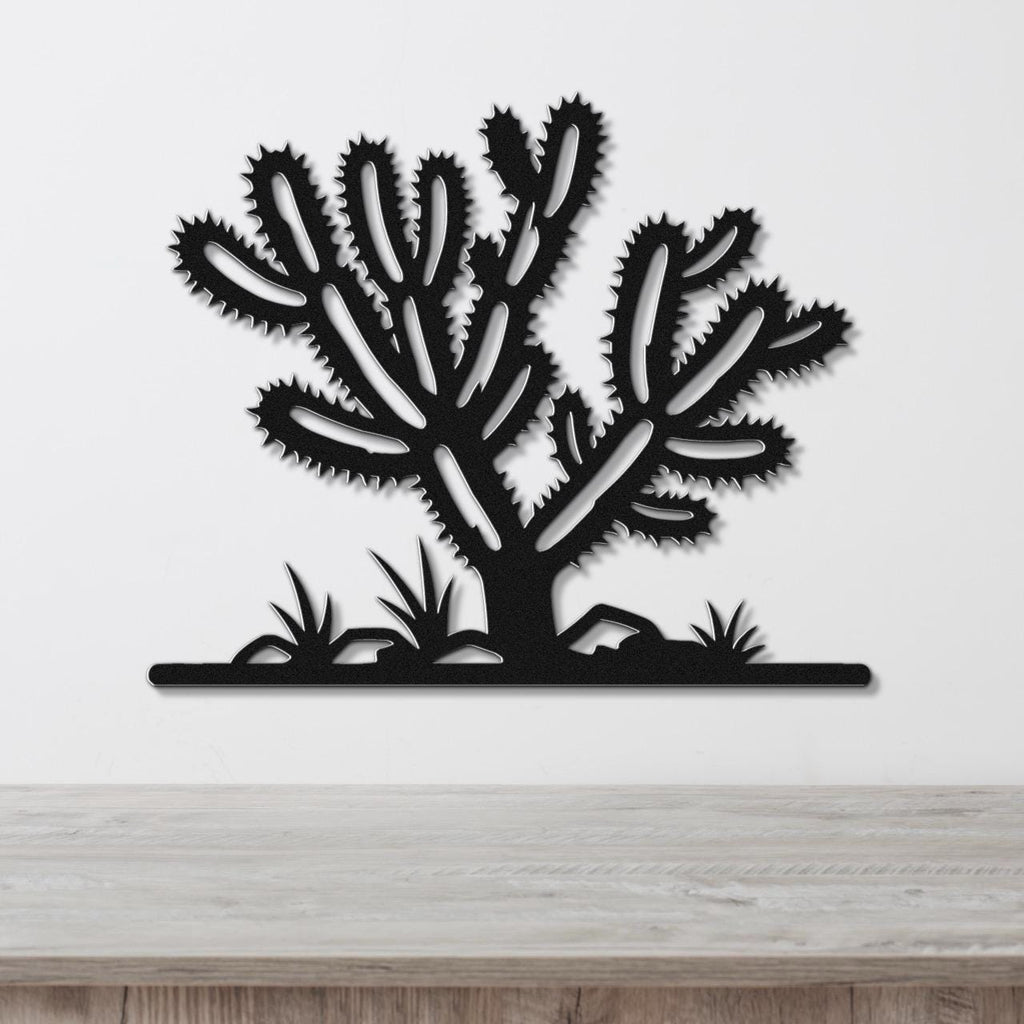 Black metal wall art cutout of a cholla cactus with spiky branches and ground-level plants, mounted on a white wall above a wooden surface.