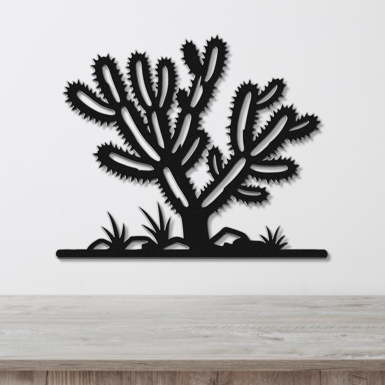 Black metal wall art cutout of a cholla cactus with spiky branches and ground-level plants, mounted on a white wall above a wooden surface.