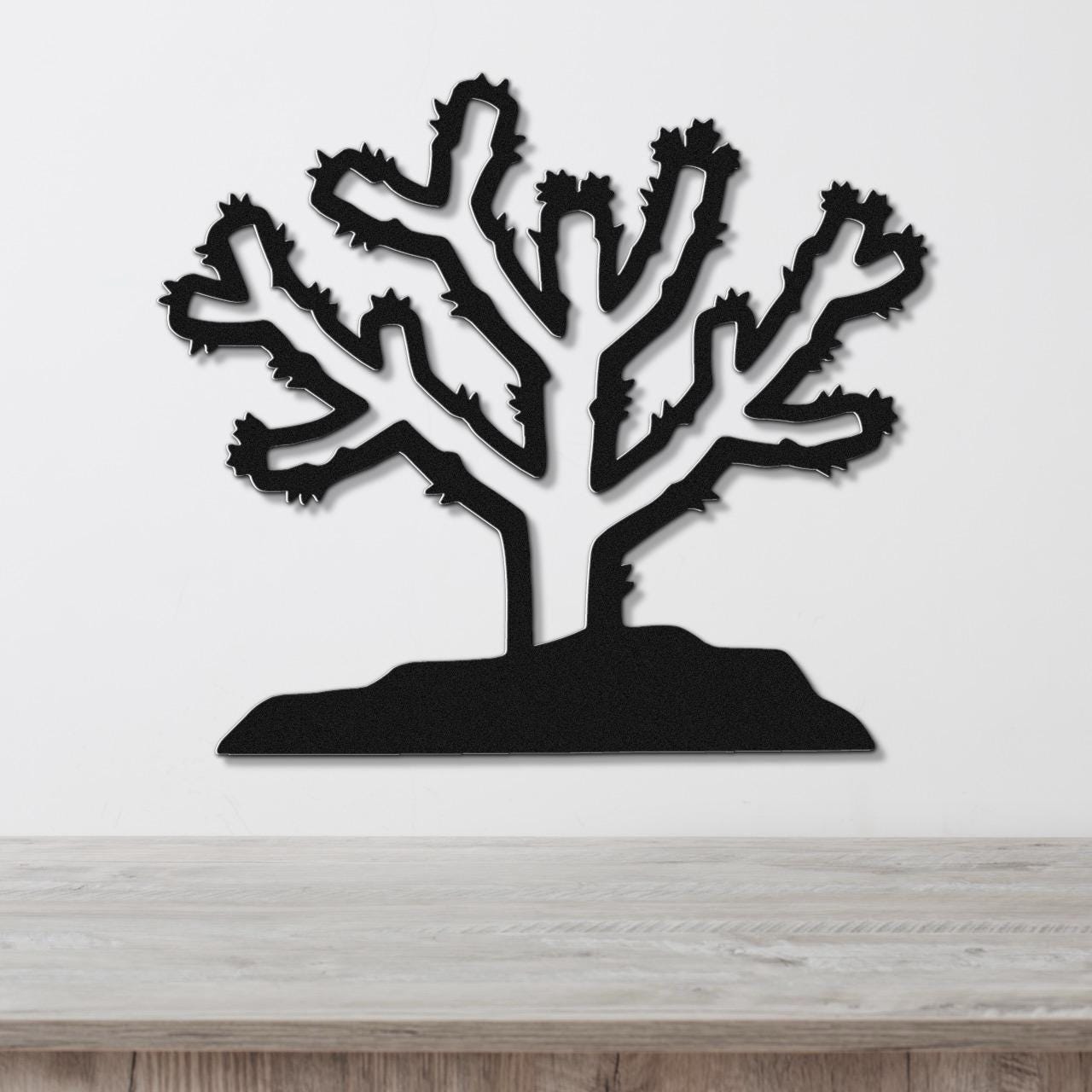 Black metal wall art cutout of a cholla cactus with branching arms and spines, mounted on a white wall above a light wood surface.