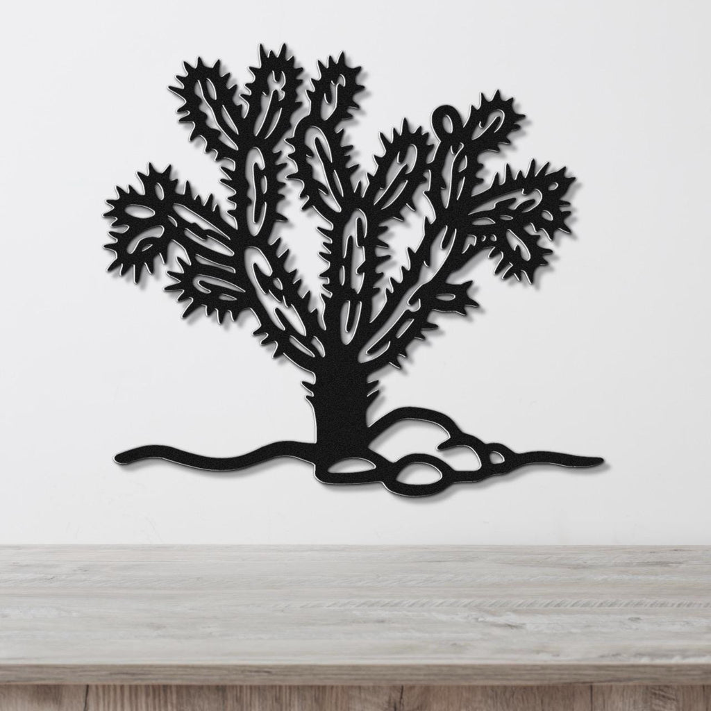 Black metal wall art cutout of a cholla cactus with detailed spines and branching arms, mounted on a white wall above a light wood surface.