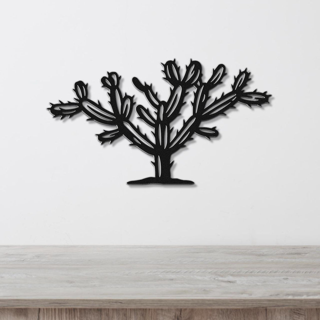 Black metal wall art cutout of a cholla cactus with branching pads and spines, mounted on a white wall above a light wood surface.