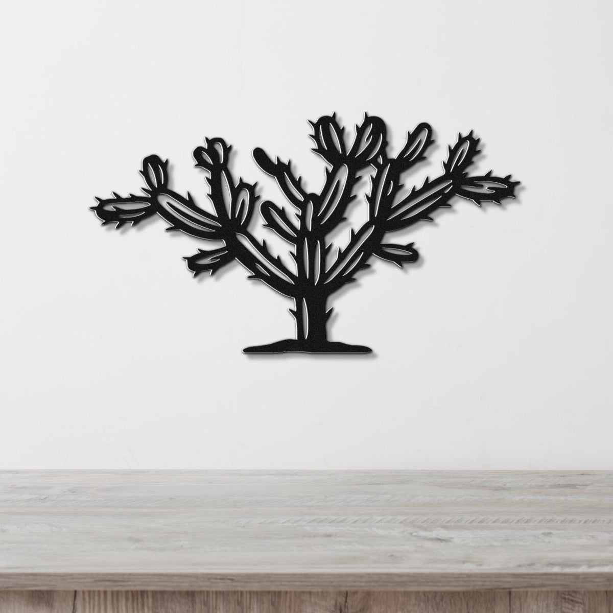 Black metal wall art cutout of a cholla cactus with branching pads and spines, mounted on a white wall above a light wood surface.