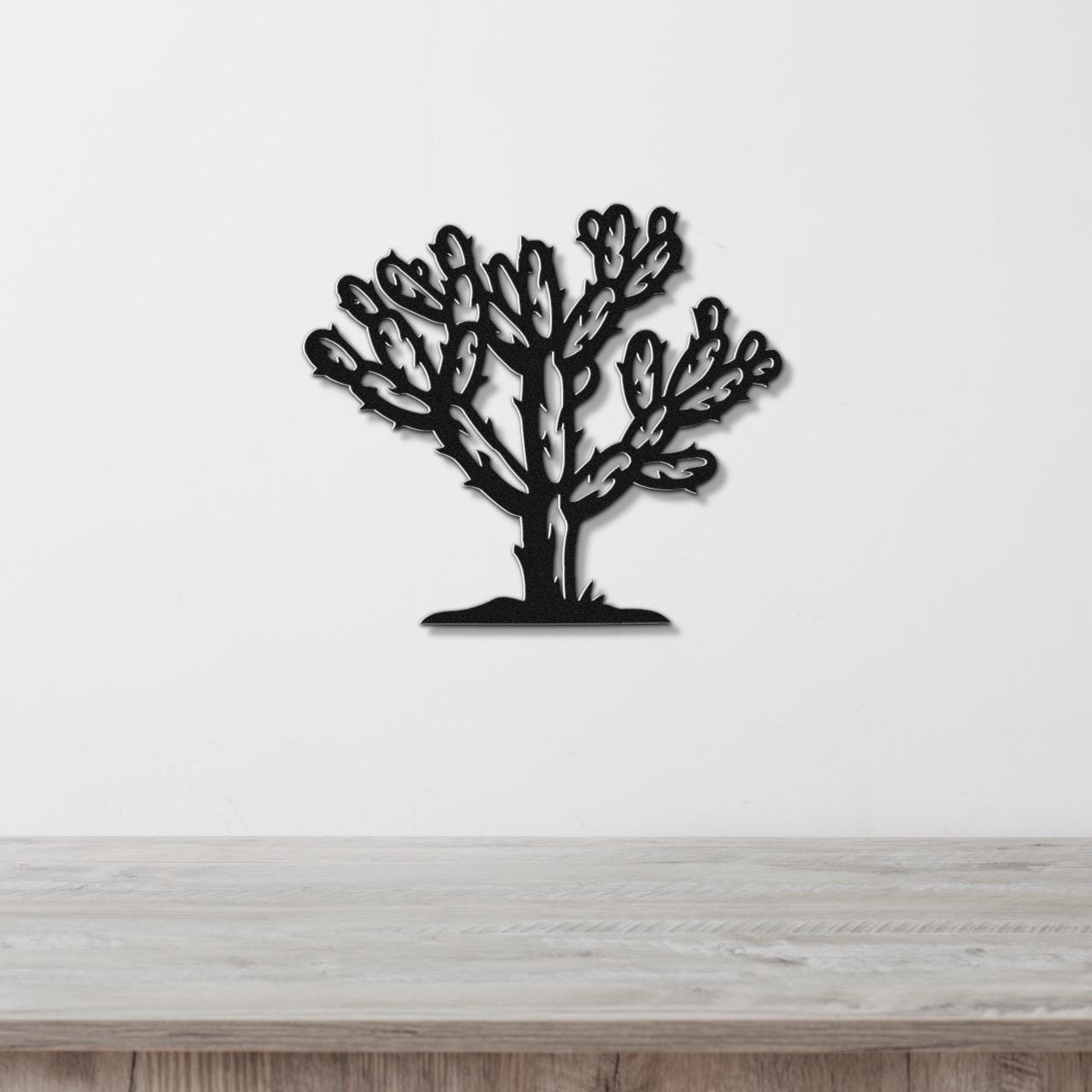 Black metal wall art cutout of a cholla cactus with detailed branching arms, mounted on a white wall above a light wood surface.