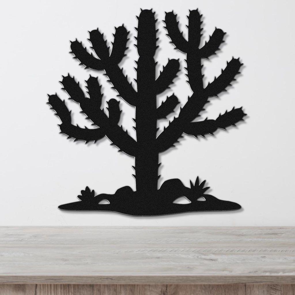 Black metal wall art cutout of a cholla cactus with spiny branches and small plants at its base, mounted on a white wall above a light wood surface.