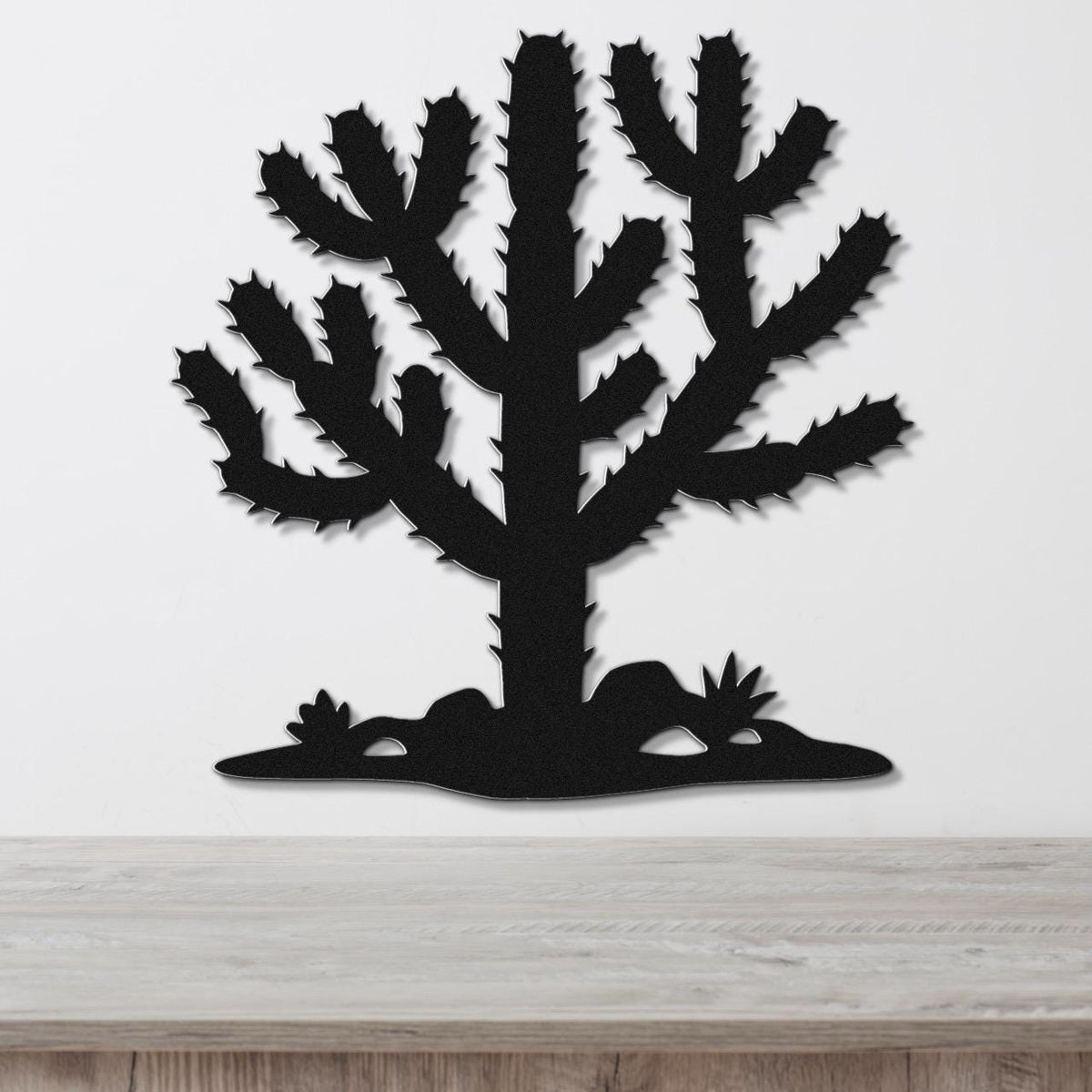Black metal wall art cutout of a cholla cactus with spiny branches and small plants at its base, mounted on a white wall above a light wood surface.