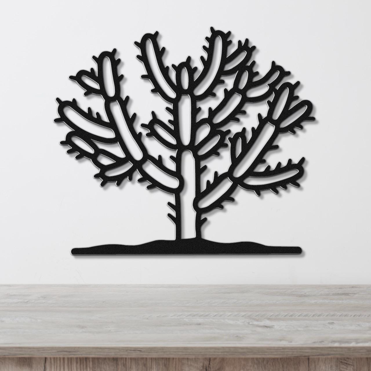 Black metal wall art cutout of a cholla cactus with branching arms and spines, mounted on a white wall above a light wood surface.