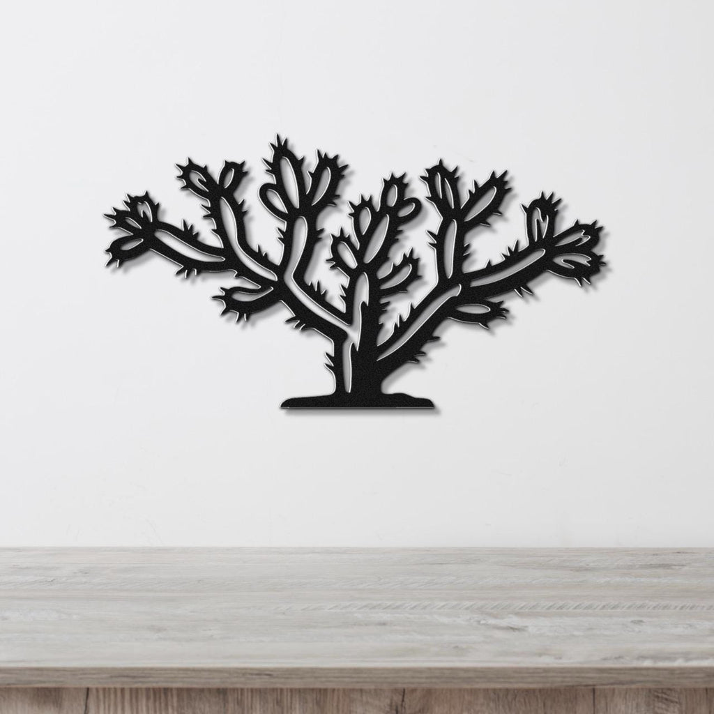 Black metal wall art in the silhouette of a cholla cactus with detailed spines and branching arms, mounted on a white wall above a light wood surface.