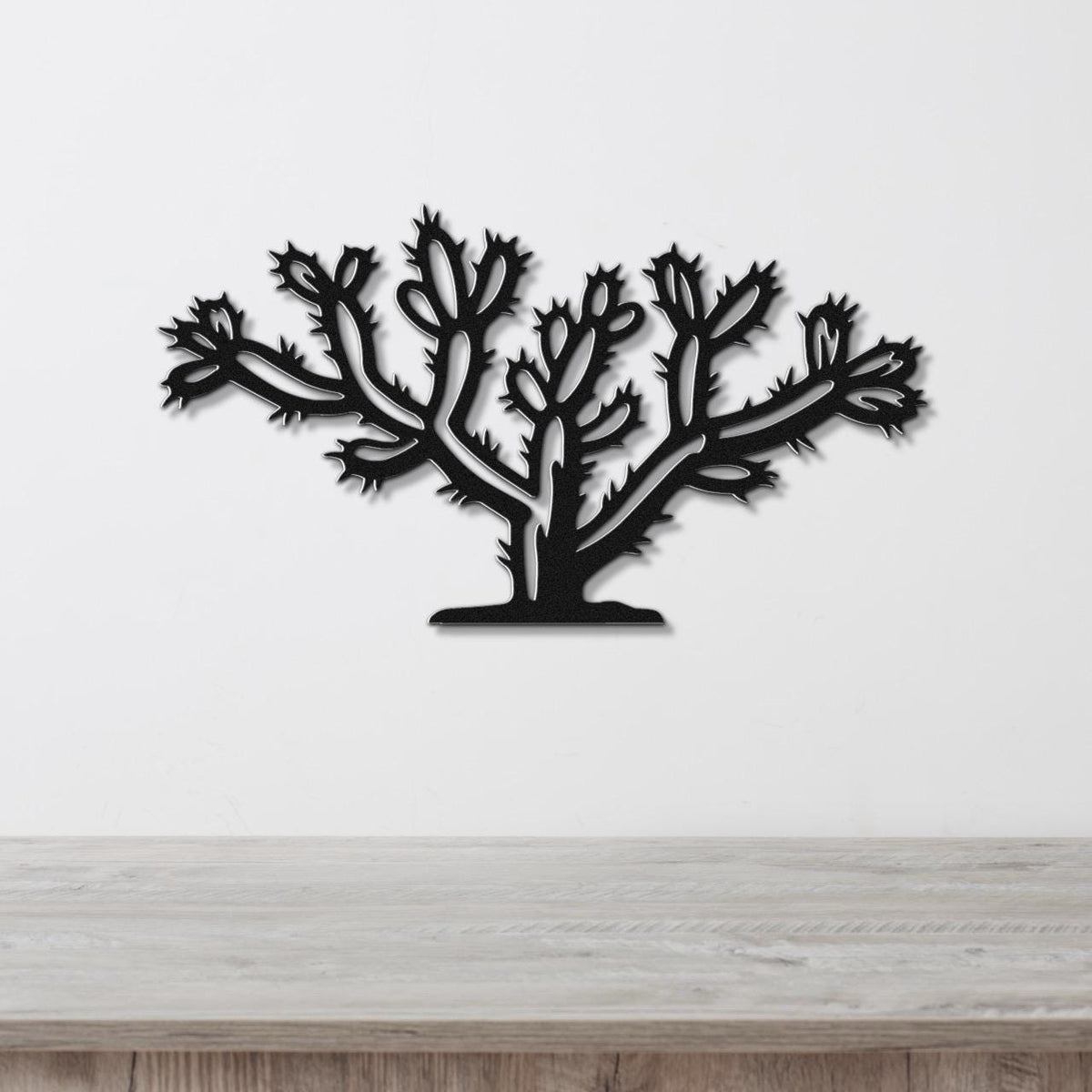 Black metal wall art in the silhouette of a cholla cactus with detailed spines and branching arms, mounted on a white wall above a light wood surface.