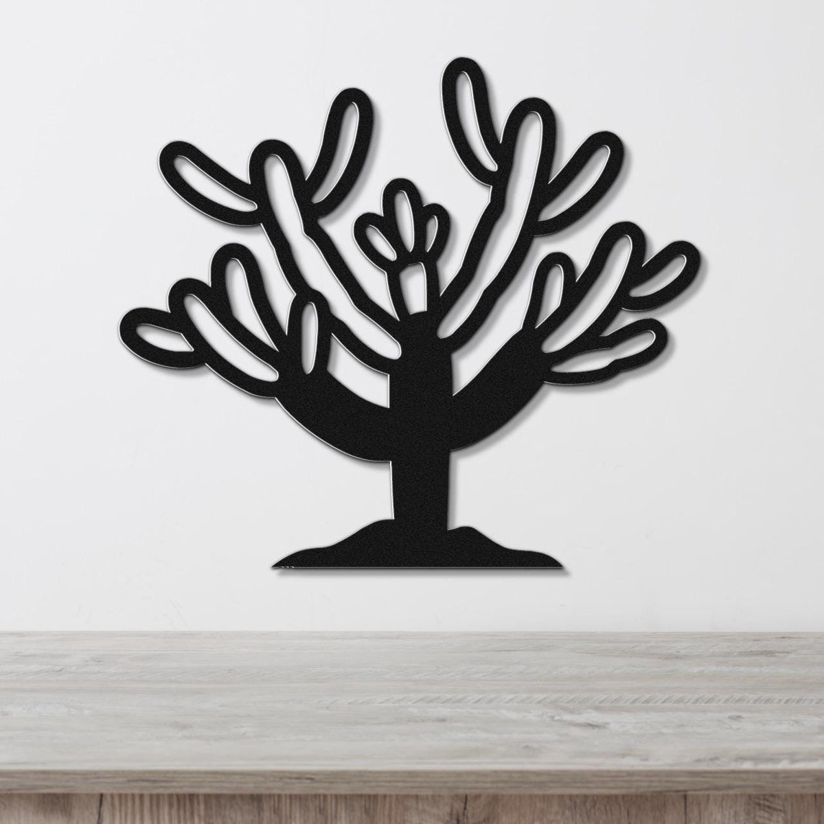 Black metal wall art cutout of a cholla cactus with branching arms and flower details, mounted on a white wall above a light wood surface.