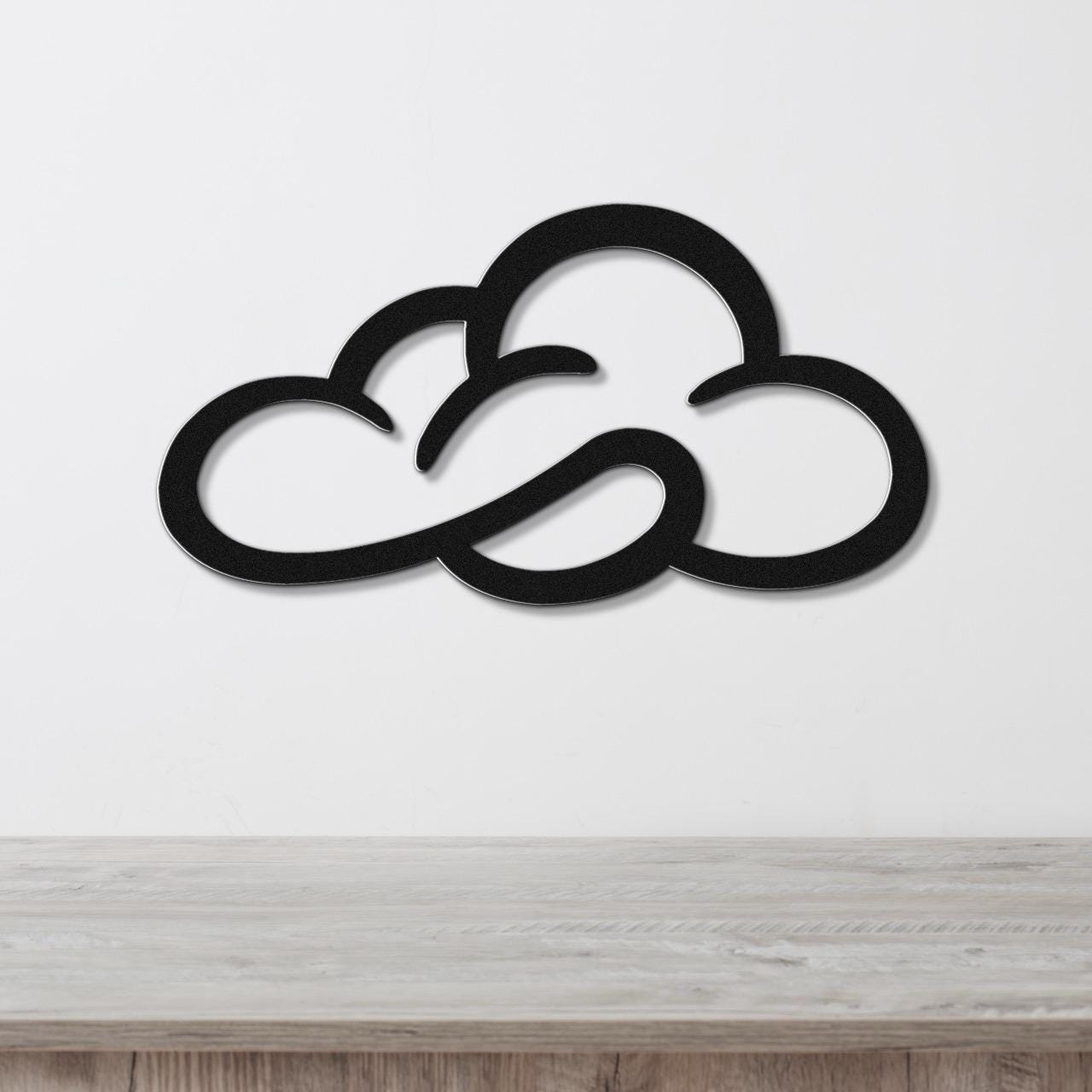 Black metal cloud wall art with open silhouette cutouts, mounted on a white wall above a light wood surface.