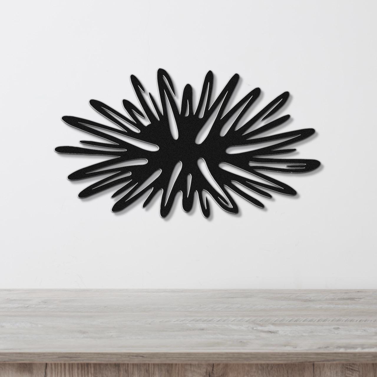 Black metal sea urchin wall art with radiating spines and internal cutouts, mounted on a white wall above a light wood surface.