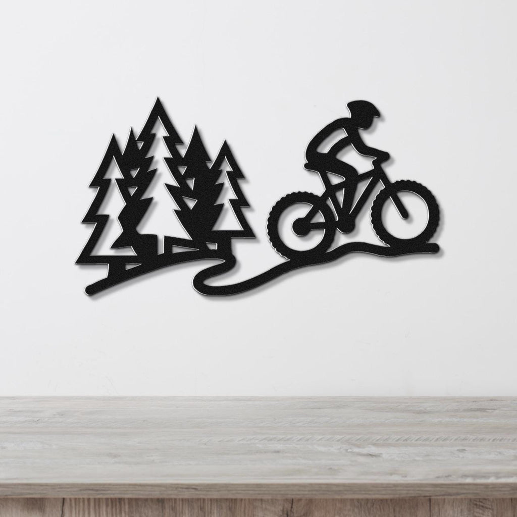 Black metal wall art cutout of a cyclist on a mountain bike trail with pine trees, mounted on a white wall above a wooden surface.