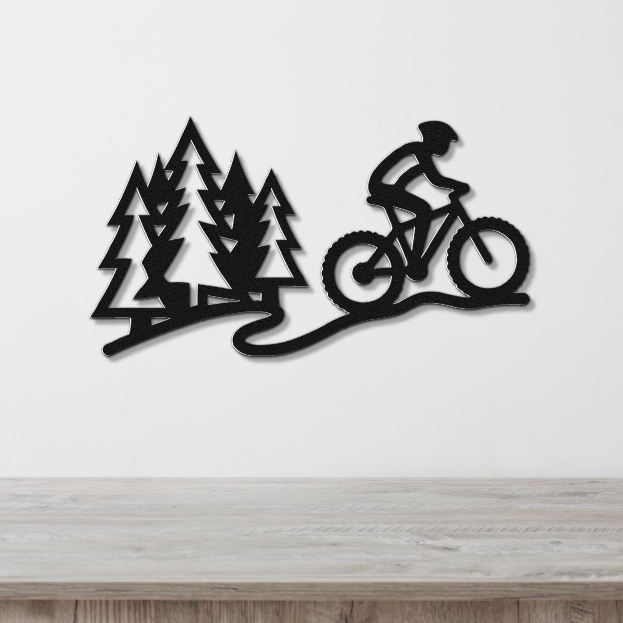 Black metal wall art cutout of a cyclist on a mountain bike trail with pine trees, mounted on a white wall above a wooden surface.