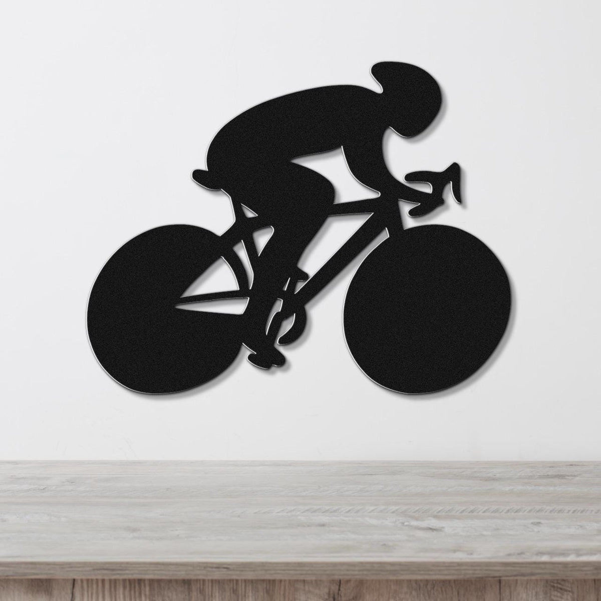 Black metal wall art featuring a bold silhouette of a cyclist in motion, mounted on a white wall above a light wood surface.