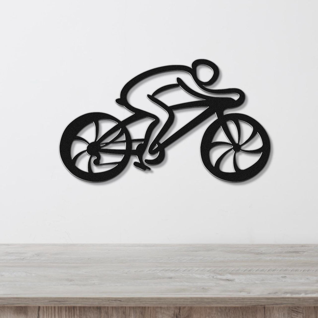 Black metal wall art cutout of a cyclist on a bike with wheel and frame cutouts, mounted on a white wall above a wooden surface.