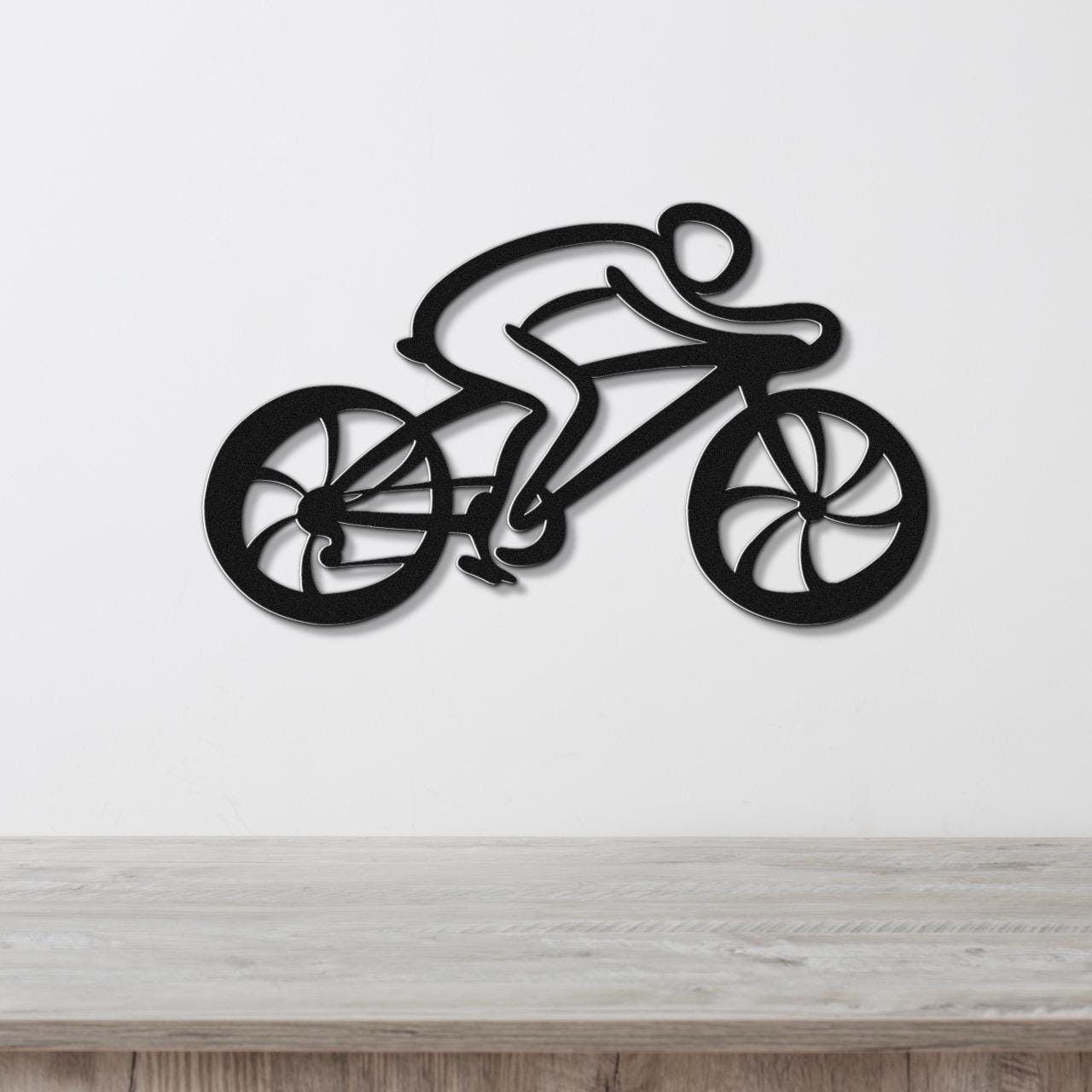 Black metal wall art cutout of a cyclist on a bike with wheel and frame cutouts, mounted on a white wall above a wooden surface.
