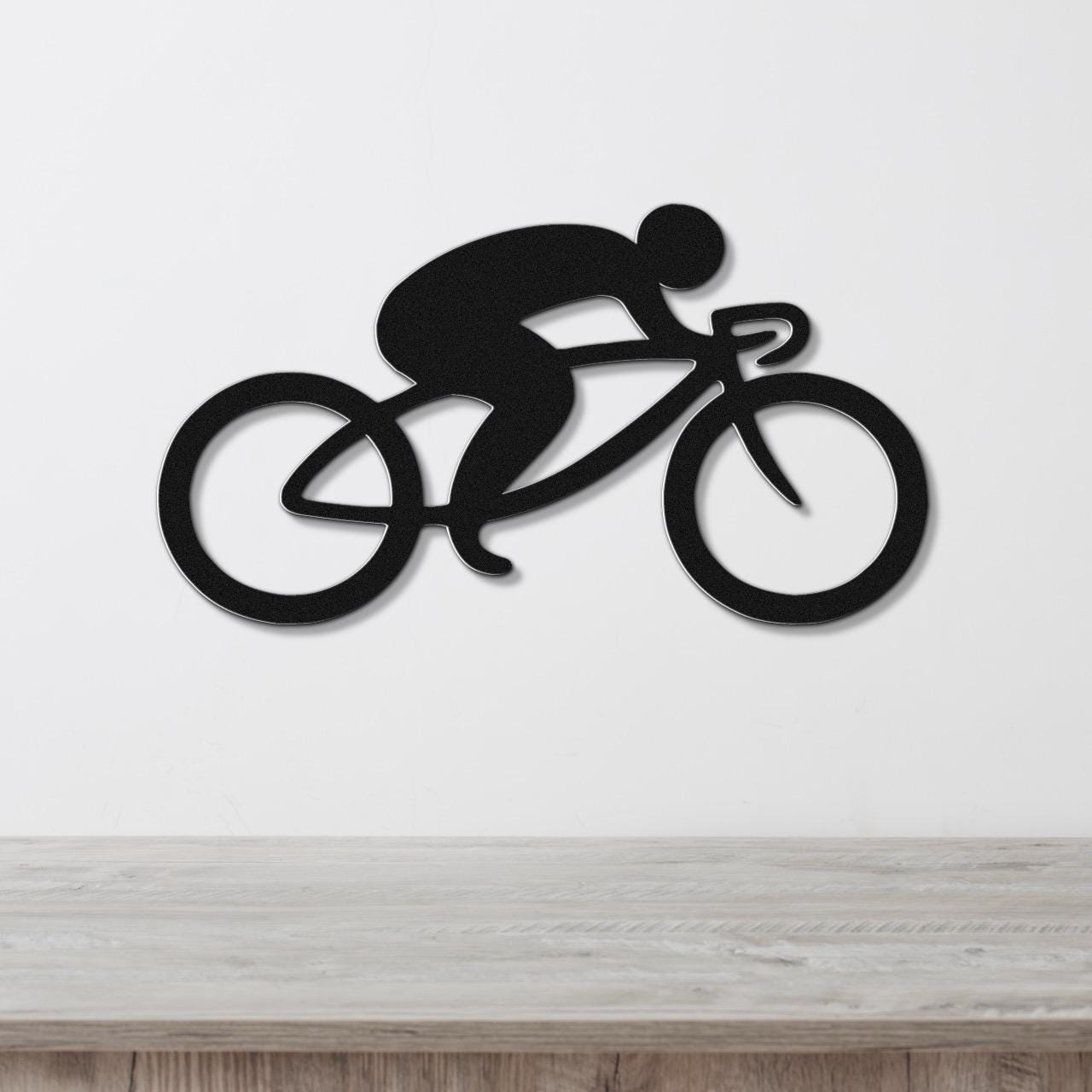 Black metal wall art featuring a minimalist cyclist silhouette in a racing pose, mounted on a white wall above a light wood surface.
