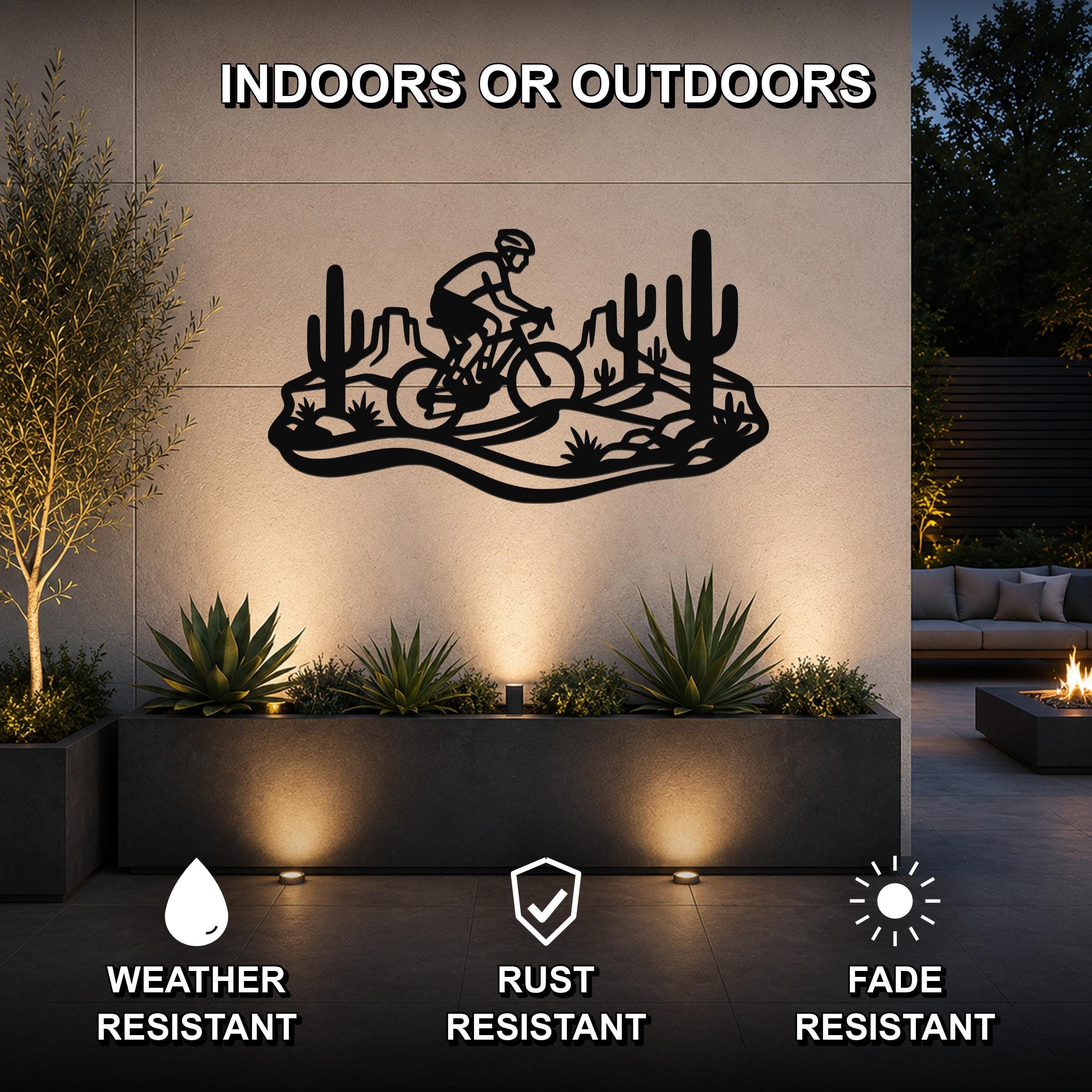 Black metal wall art featuring a cyclist silhouette riding through a Sonoran desert scene with cacti and rock formations, mounted on an outdoor wall above a planter in a patio setting.