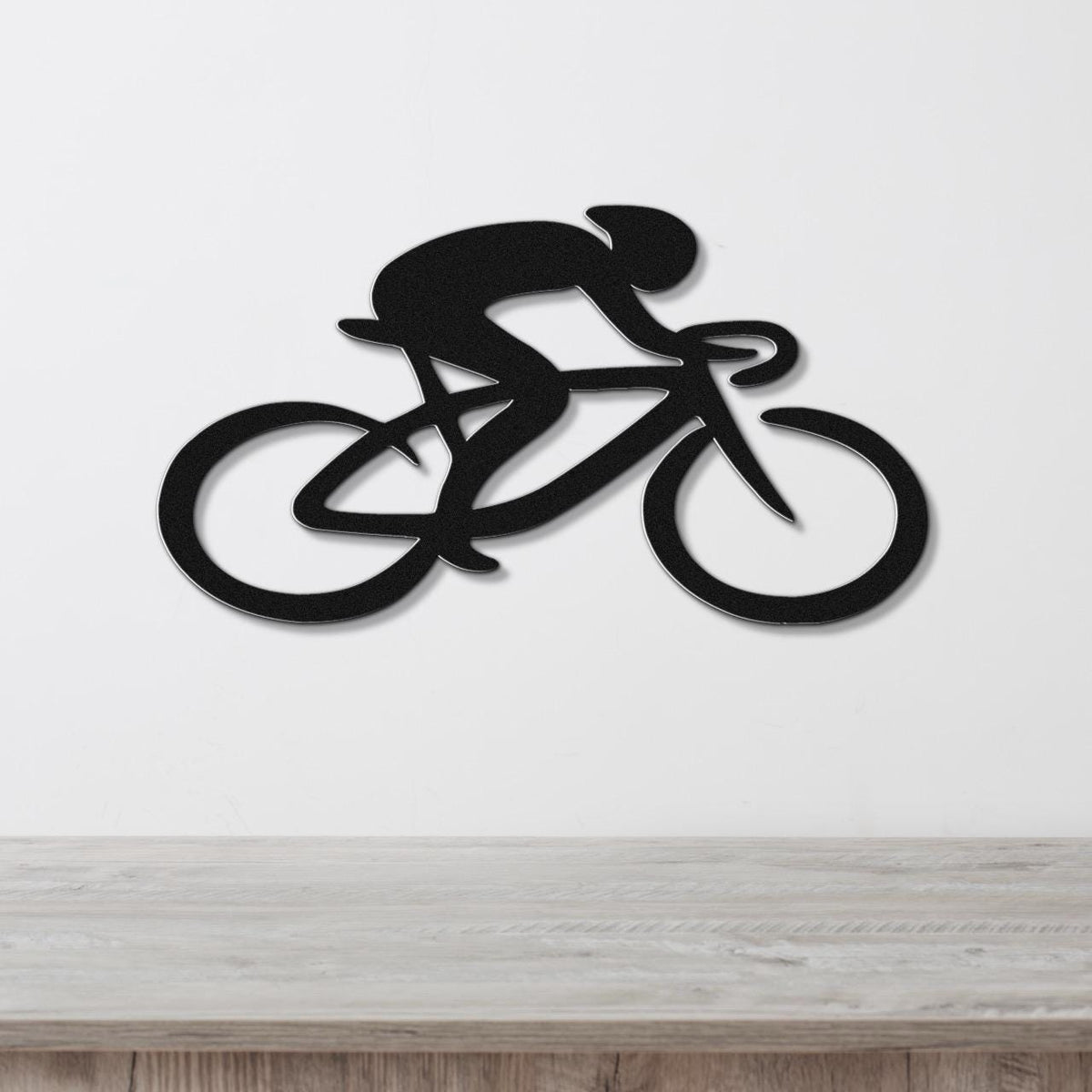 Black metal wall art featuring a cyclist in a dynamic silhouette with cutout wheels and frame, mounted on a white wall above a wooden surface.