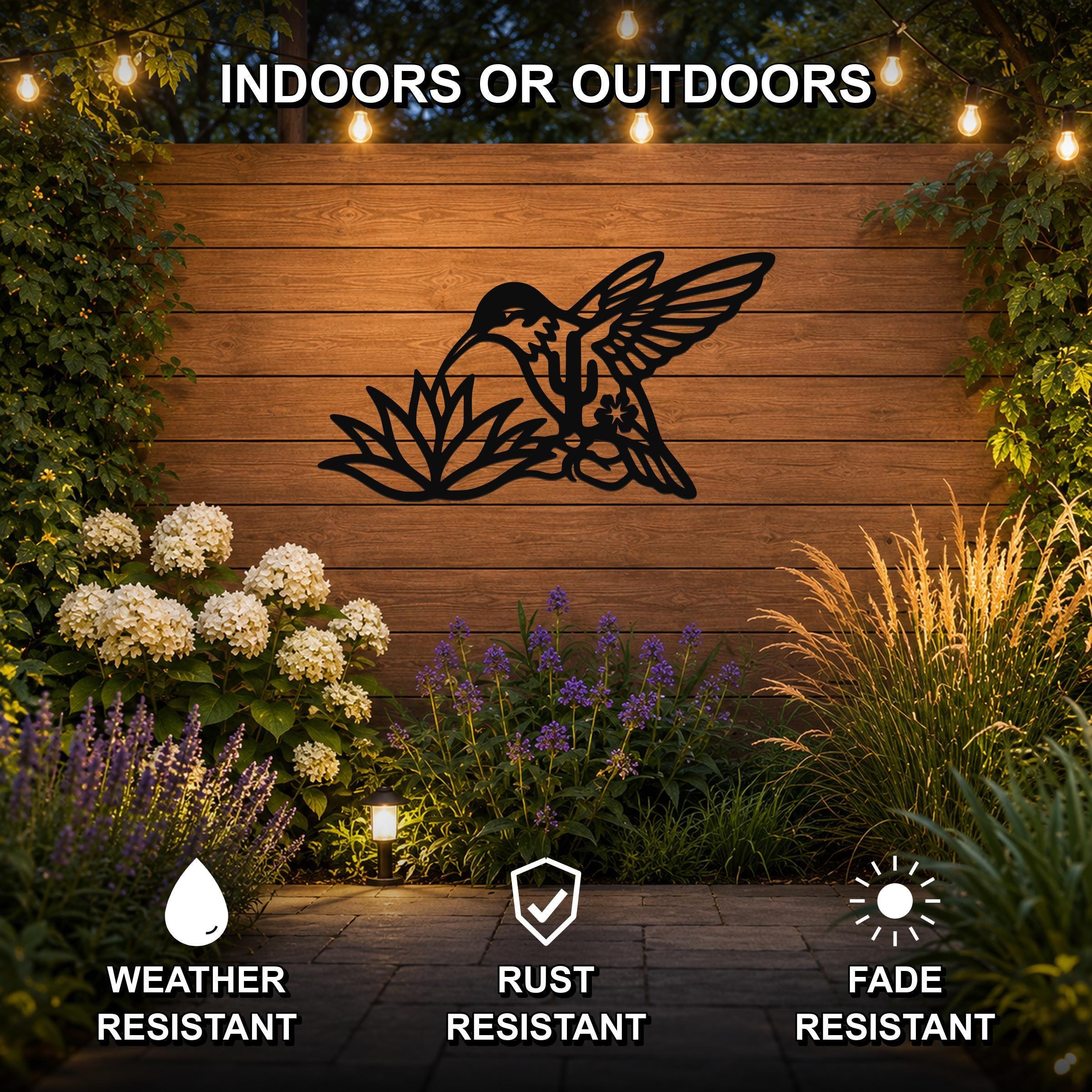 Black metal hummingbird wall art with flower and leaf cutouts, mounted on a wooden outdoor wall above a garden with string lights and plants.