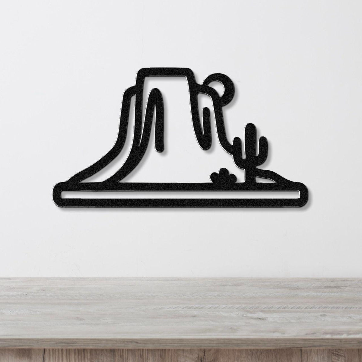 Black metal wall art cutout of a desert mesa and cactus, mounted on a white wall above a wooden surface.