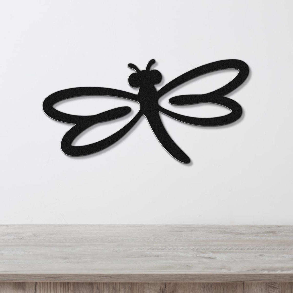 Black metal dragonfly wall art with cutout wings and body, mounted on a white wall above a light wood surface.