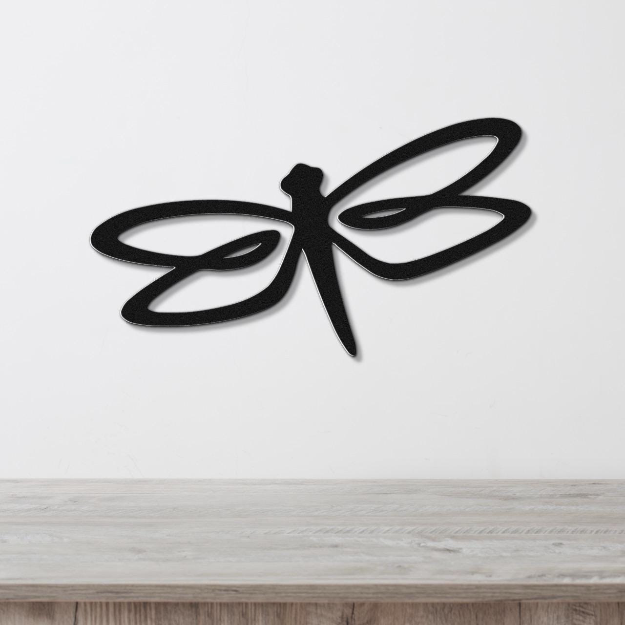 Black metal dragonfly wall art with flowing wing cutouts, mounted on a white wall above a light wood surface.