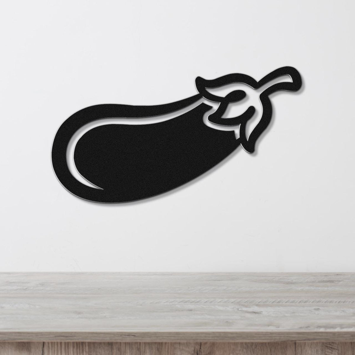 Black metal eggplant wall art with a cutout stem and leaf, mounted on a white wall above a light wood countertop.