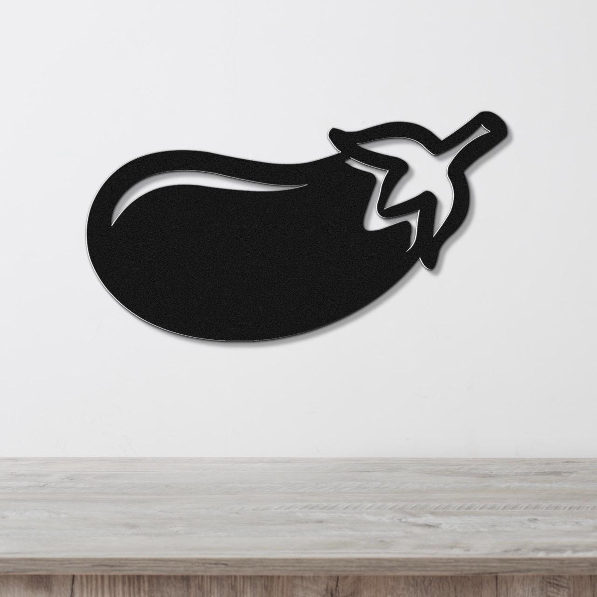 Black metal eggplant wall art with a cutout stem and leaf, mounted on a white wall above a light wood countertop.