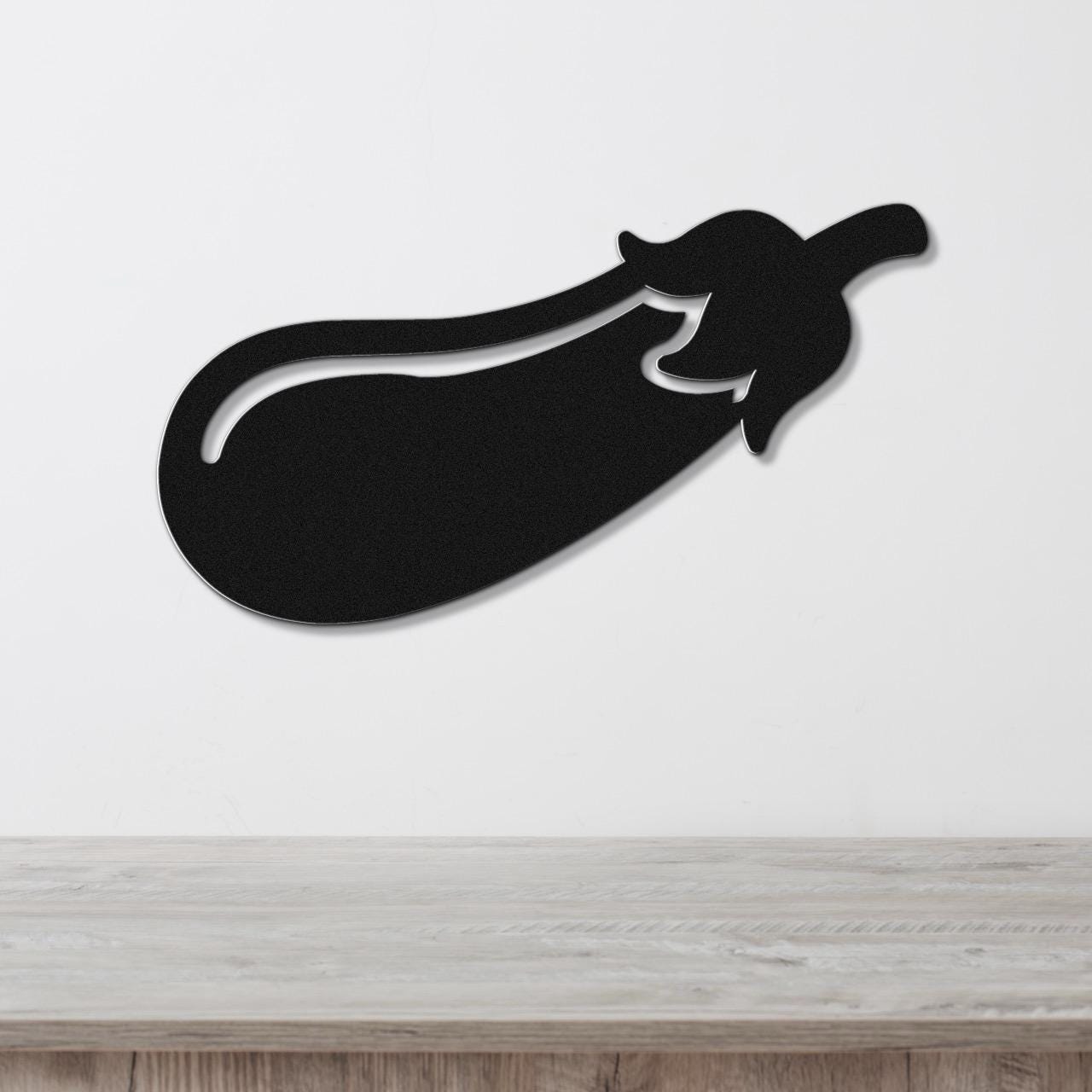 Black metal eggplant wall sculpture with a cutout stem and leaf detail, mounted on a white wall above a light wood countertop.