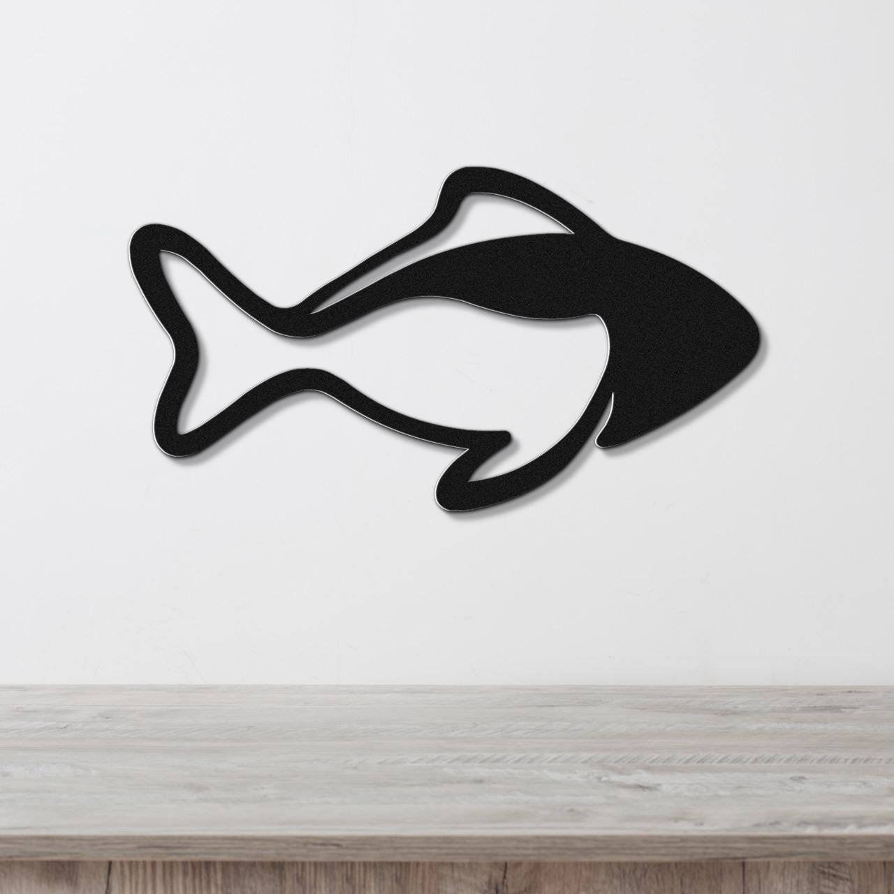 Black metal fish wall art with bold line silhouette cutout, mounted on a white wall above a light wood surface.