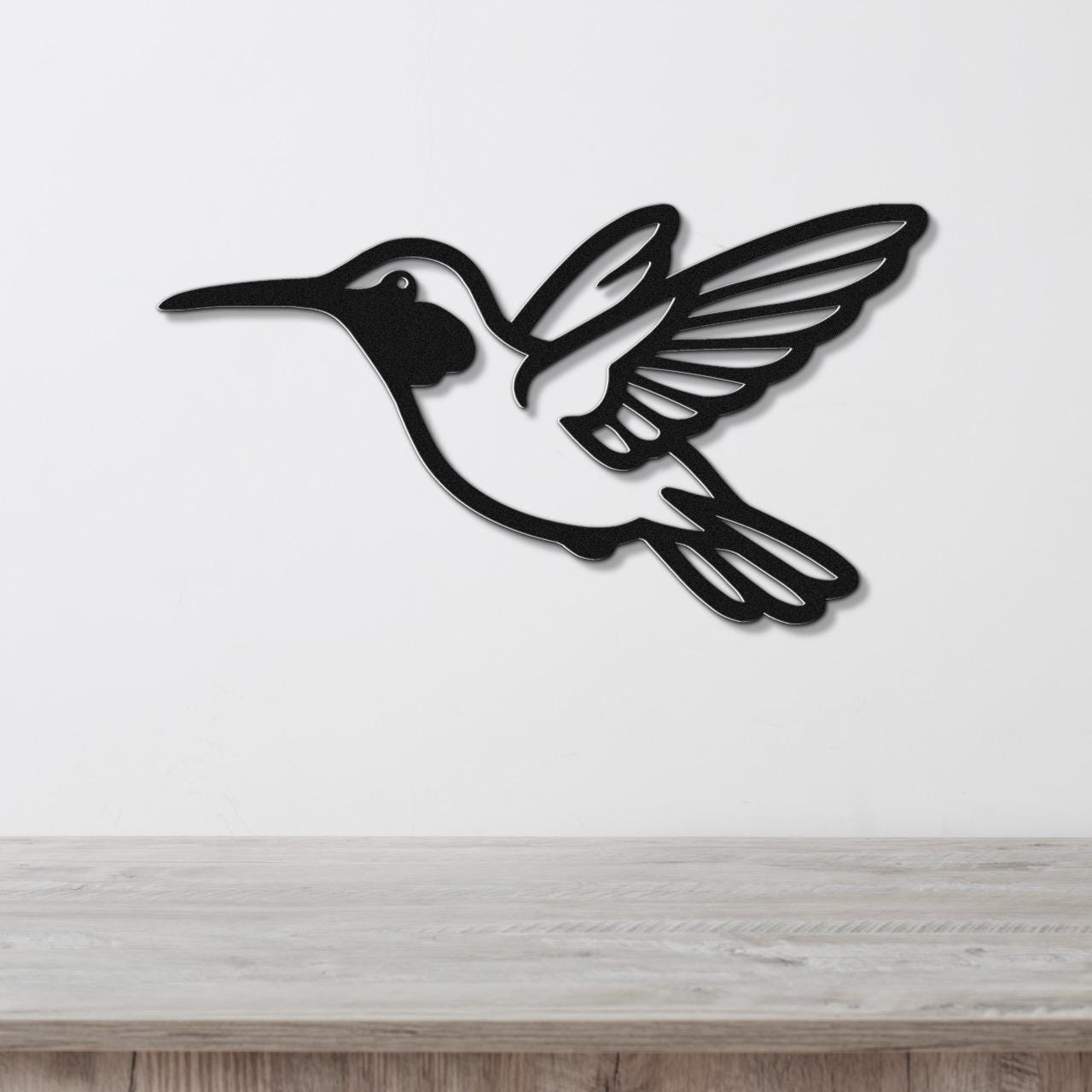 Black metal hummingbird wall art with wing and tail cutouts, mounted on a white wall above a light wood surface.