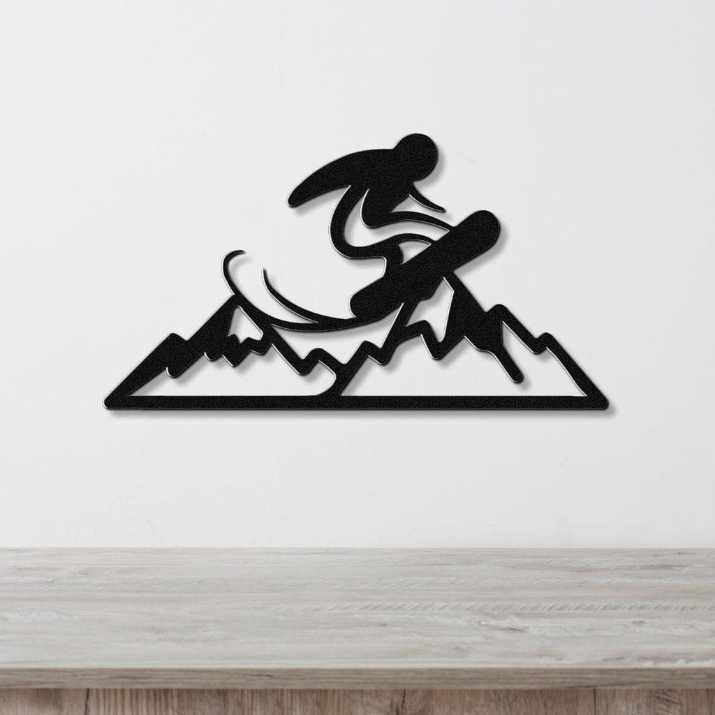 Black metal wall art featuring a snowboarder silhouette mid-jump over mountain peaks, mounted on a white wall above a wooden surface.