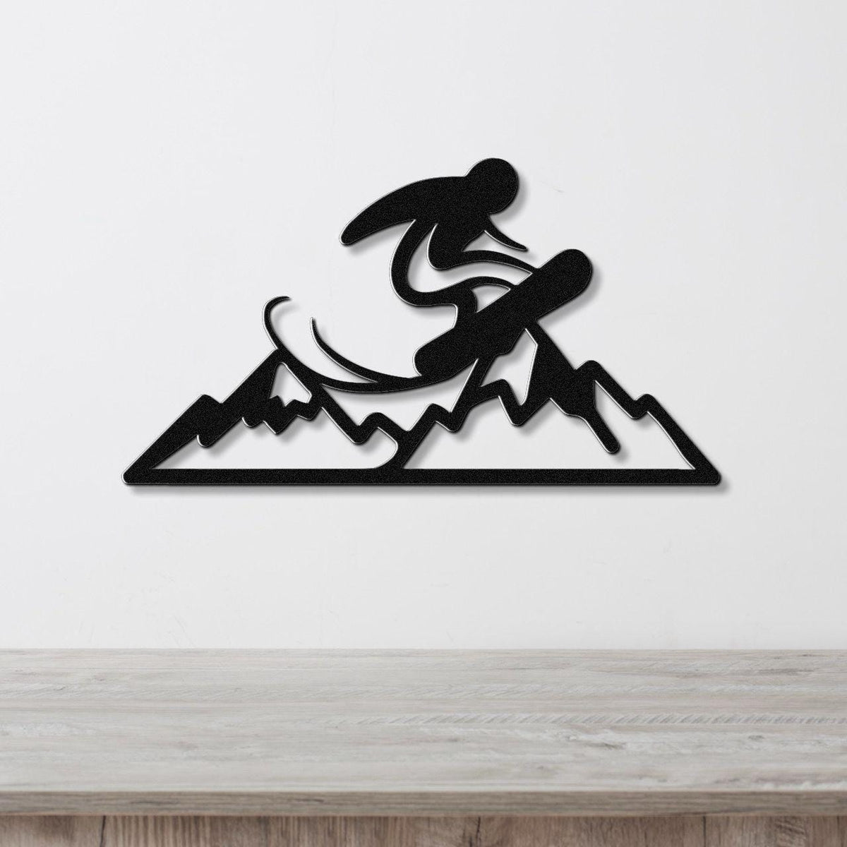 Black metal wall art featuring a snowboarder silhouette mid-jump over mountain peaks, mounted on a white wall above a wooden surface.
