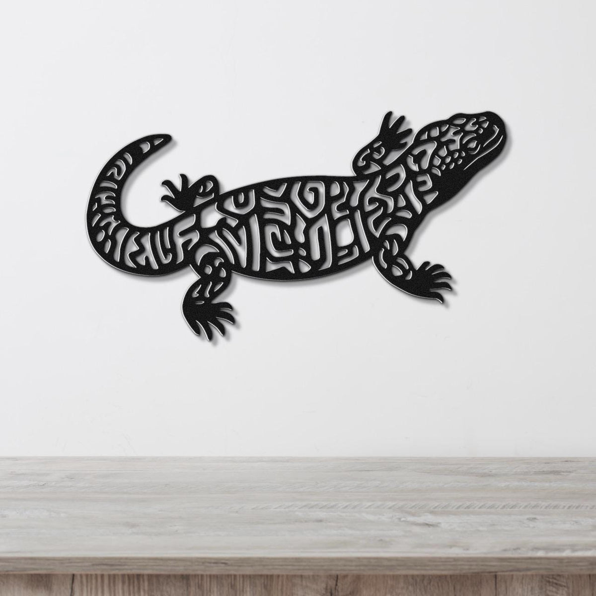 Black metal wall art of a Gila monster with intricate cutout patterns, mounted on a white wall above a wooden surface.