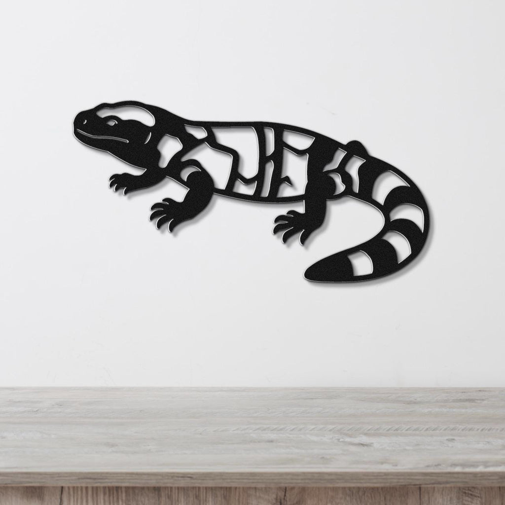 Black metal Gila monster wall art with internal cutout patterns, mounted on a white wall above a light wood surface.
