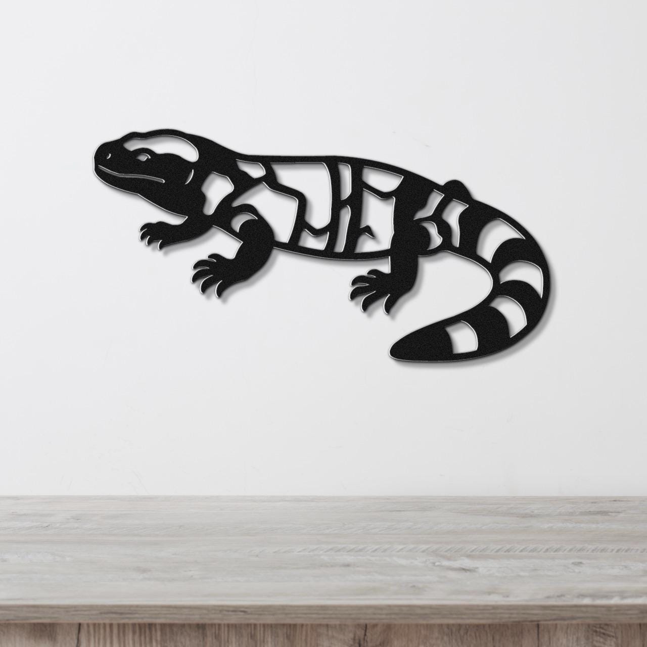 Black metal Gila monster wall art with internal cutout patterns, mounted on a white wall above a light wood surface.