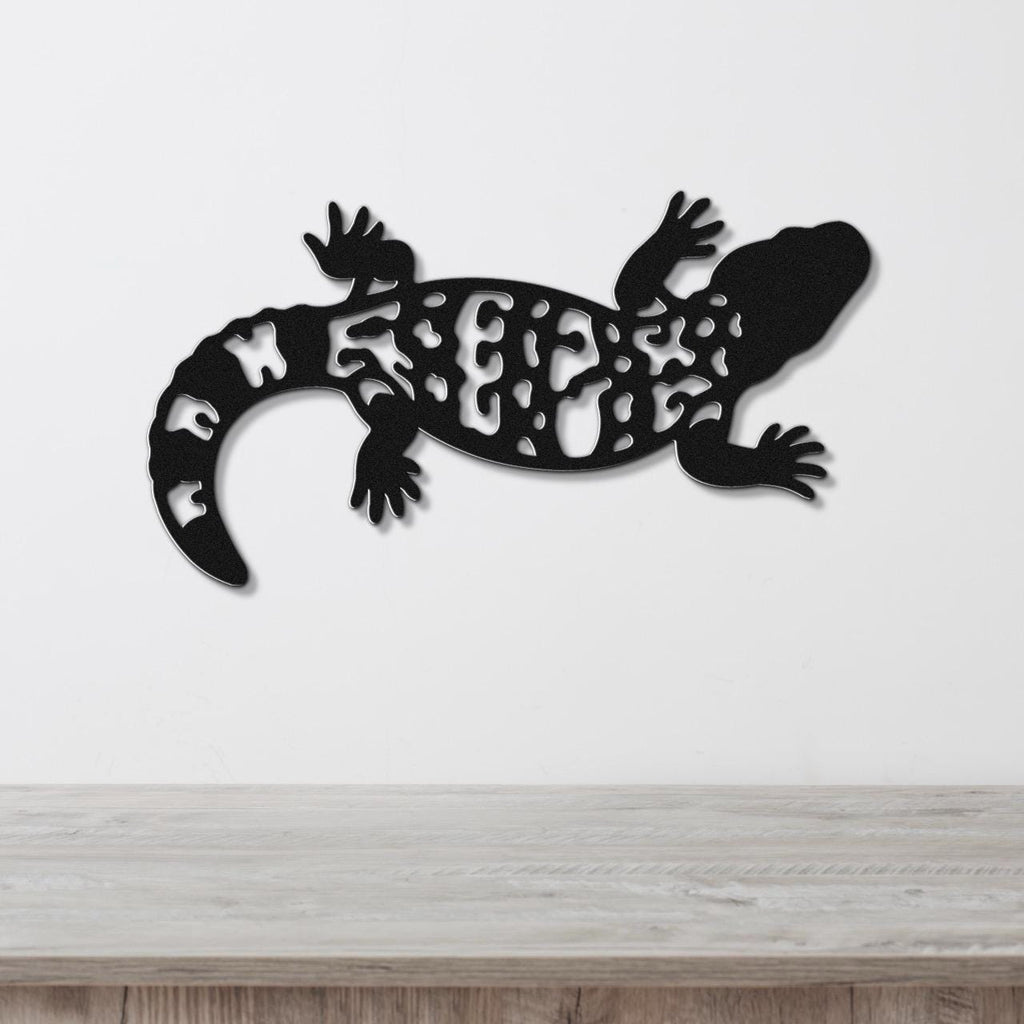 Black metal wall art of a Gila monster with intricate cutout patterns on its body, mounted on a white wall above a light wood surface.