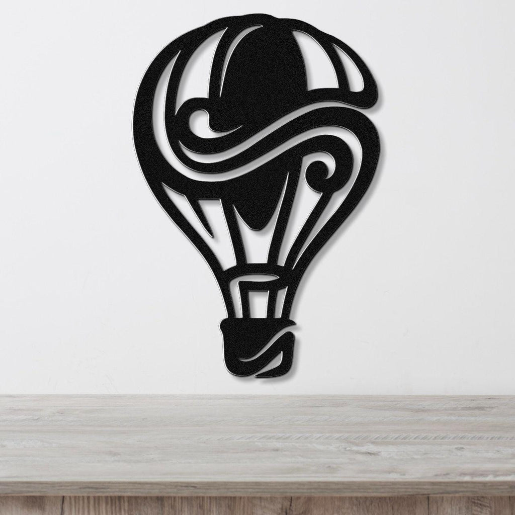 Black metal wall art in a bold hot air balloon silhouette with swirling cutout details, mounted on a white wall above a wooden surface.