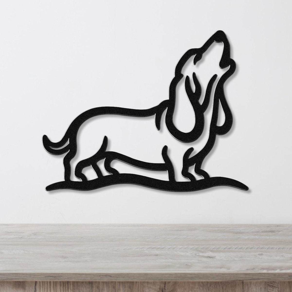 Black metal wall art of a howling basset hound in a bold silhouette, mounted on a white wall above a light wood surface.