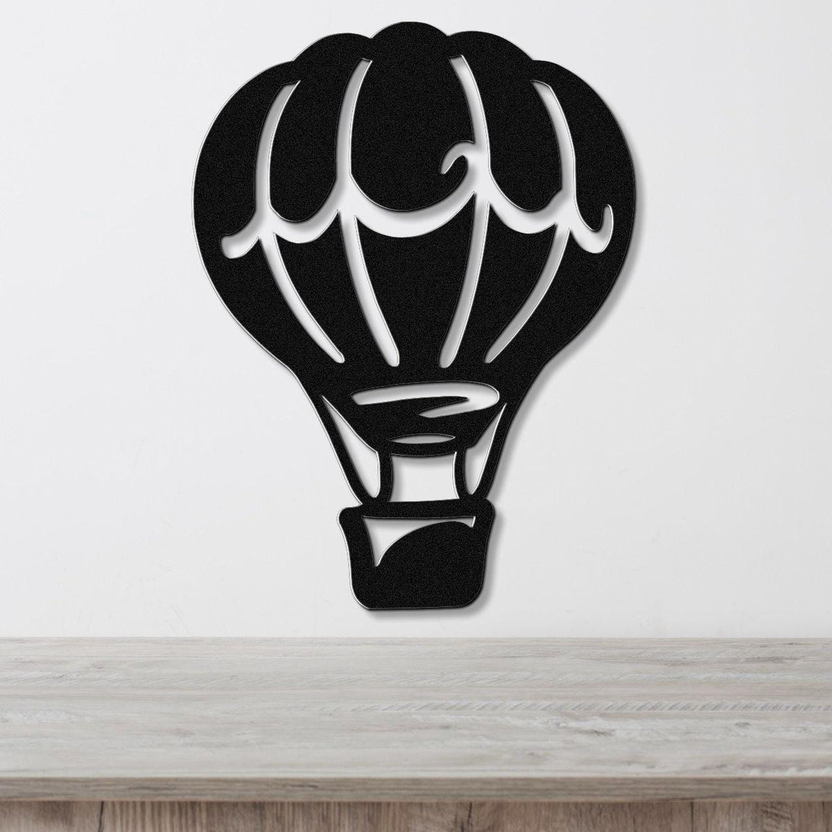 Black metal wall art in the silhouette of a hot air balloon with internal cutout details, mounted on a white wall above a light wood surface.