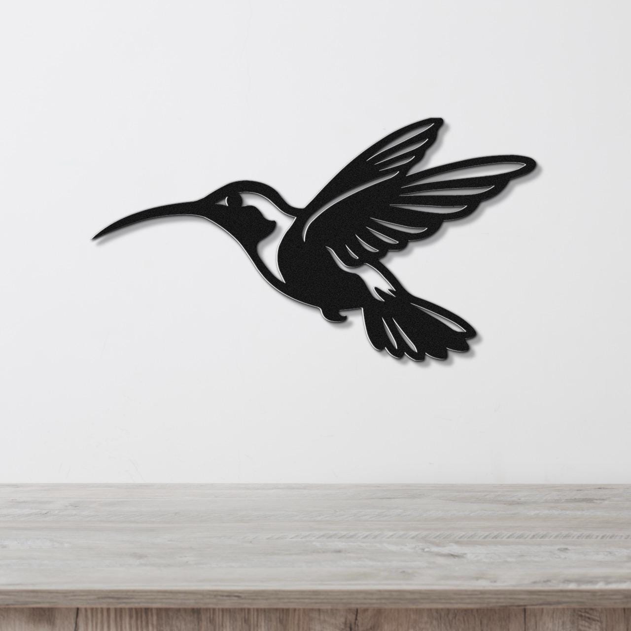 Black metal hummingbird wall art with wing and tail cutouts, mounted on a white wall above a light wood surface.