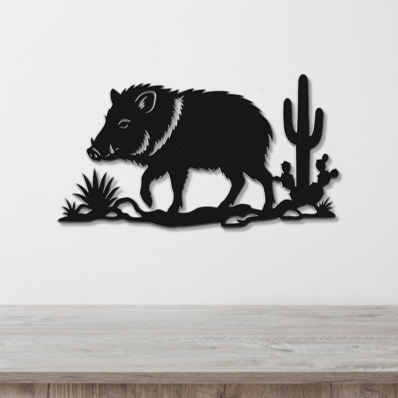 Black metal wall art featuring a javelina silhouette with cactus and desert plants, mounted on a white wall above a wooden surface.