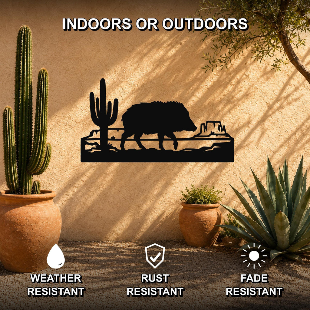 Black metal wall art featuring a javelina silhouette walking past a saguaro cactus and canyon rock formation, mounted on an outdoor stucco wall with potted cacti nearby.