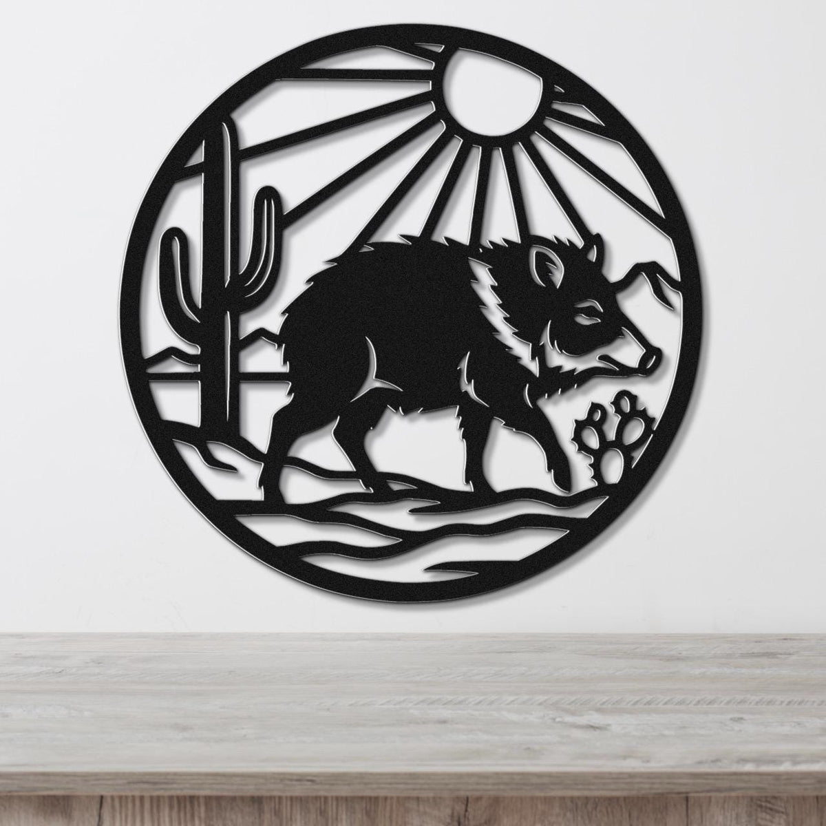 Black metal circular wall art featuring a javelina silhouette, cactus, sun rays, and paw prints, mounted on a white wall above a wooden surface.