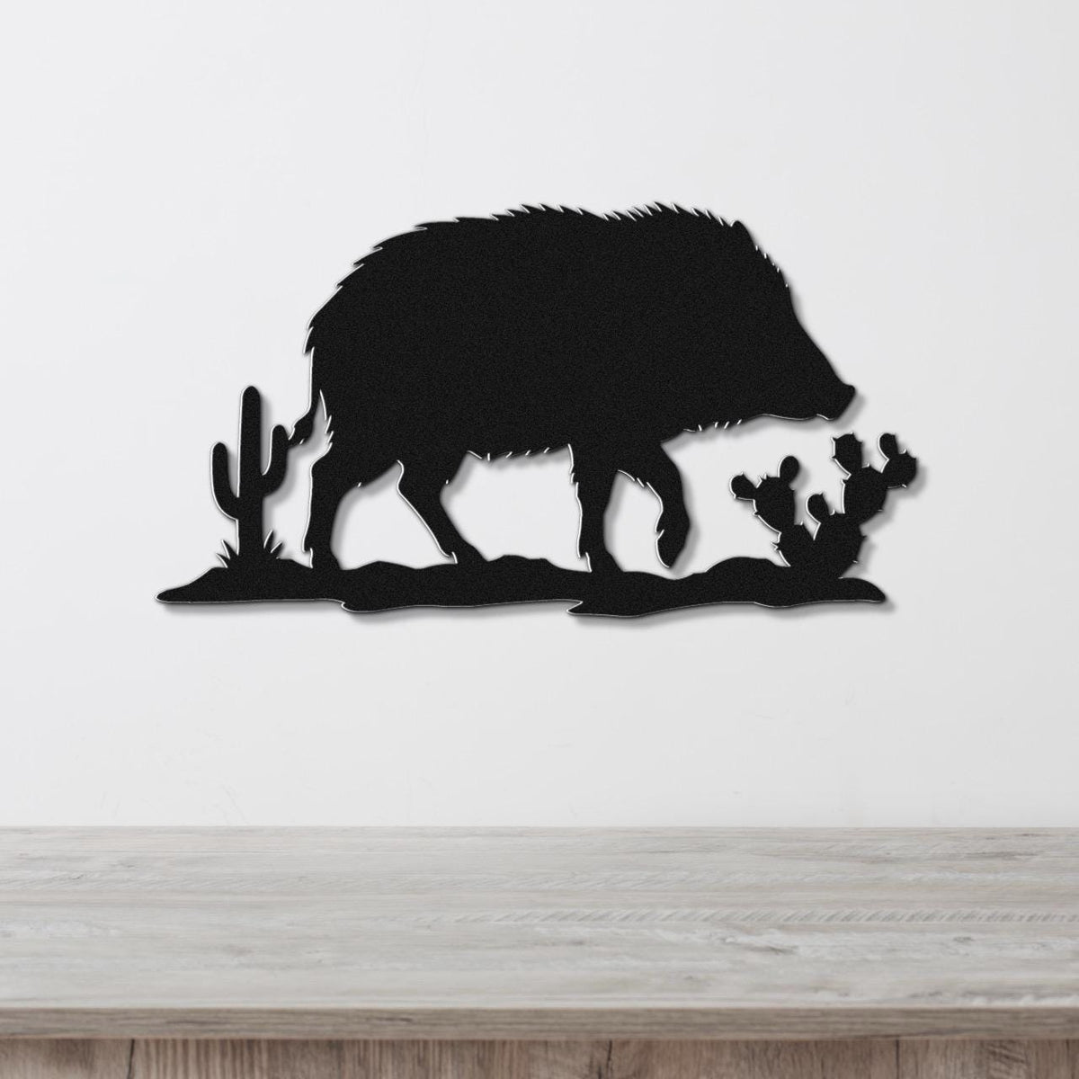 Black metal wall art silhouette of a javelina with cacti, mounted on a white wall above a wooden surface.