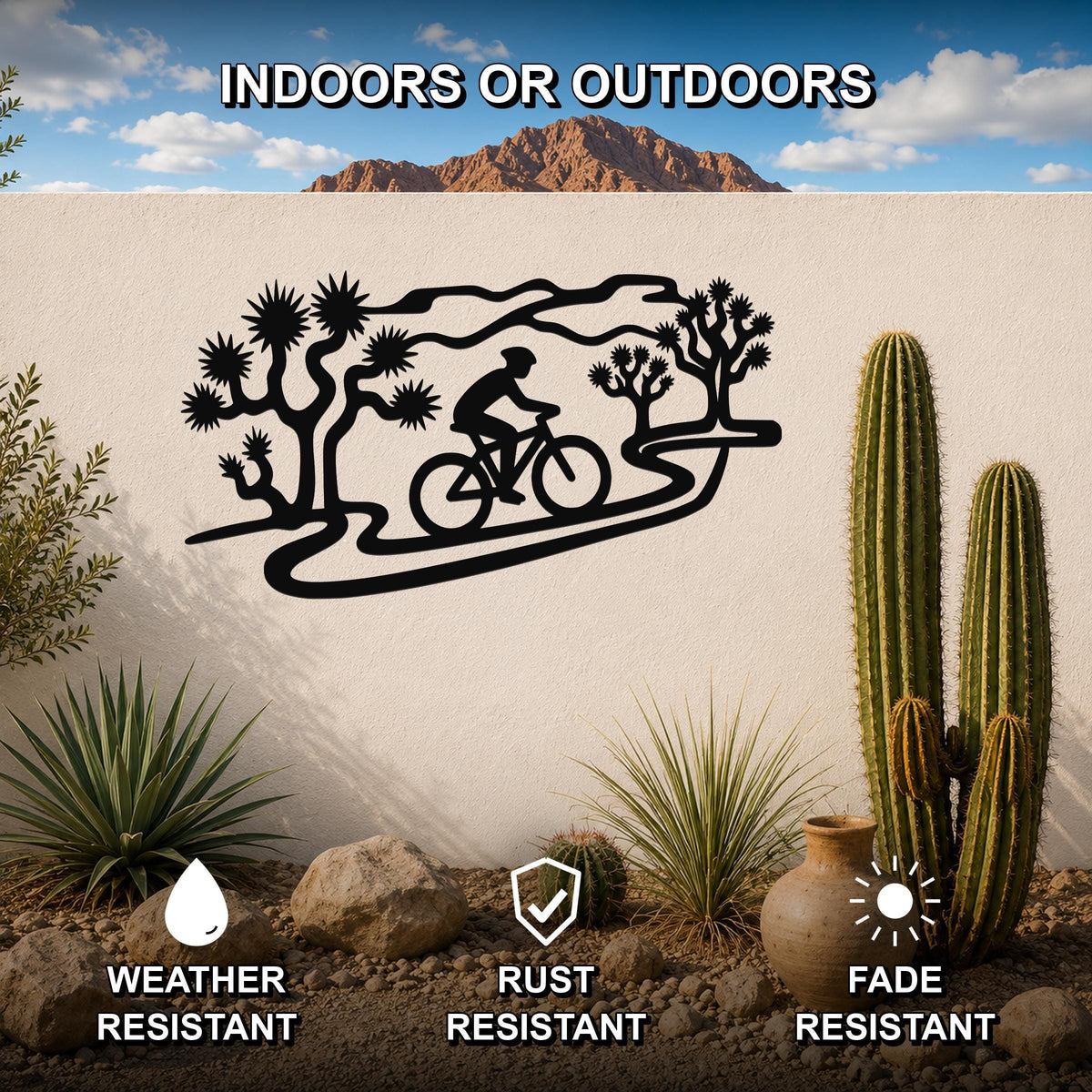 Black metal wall art featuring a cyclist silhouette on a winding path surrounded by Joshua trees, mounted on an outdoor wall in a desert landscape with cacti and rocks.