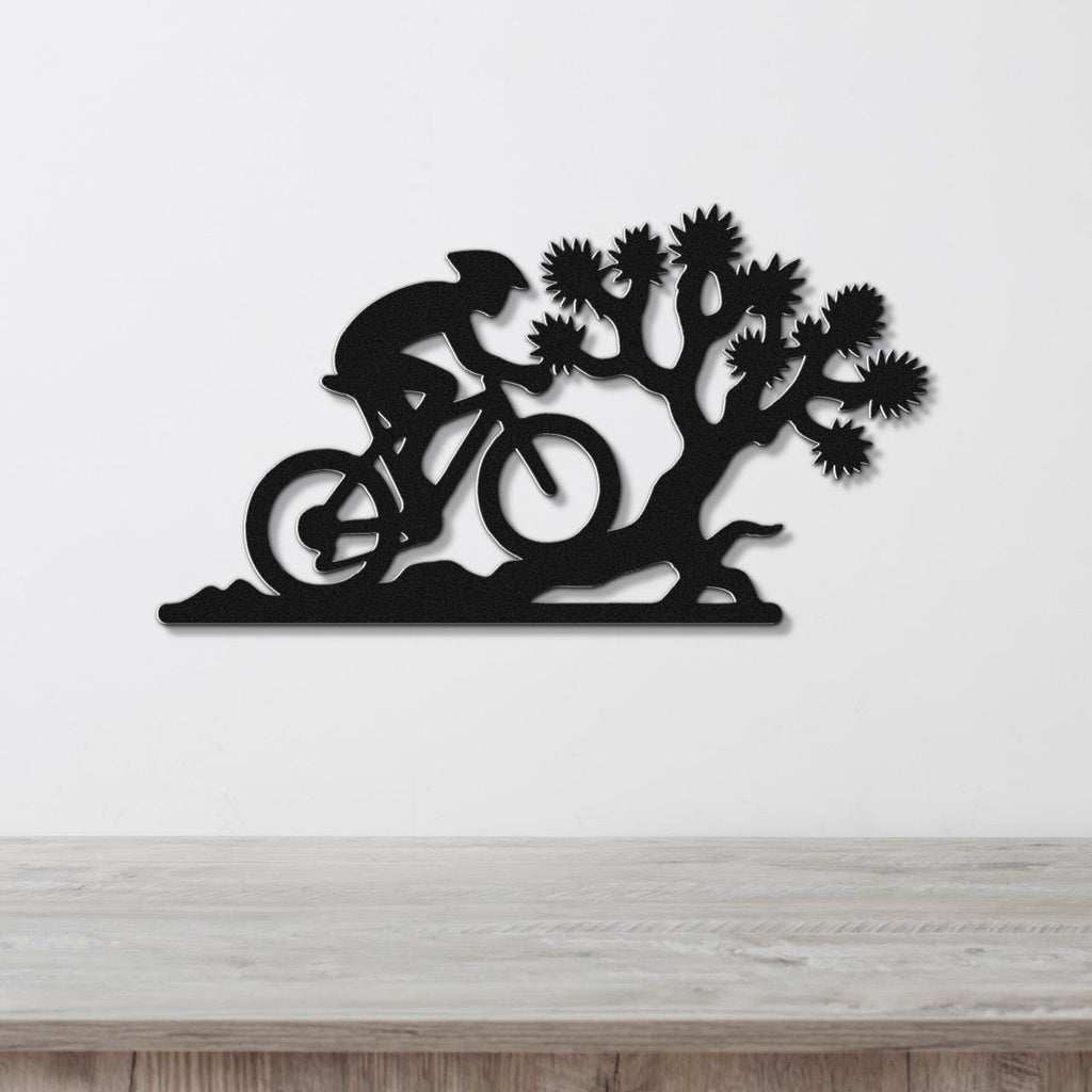 Black metal wall art cutout of a cyclist on a mountain bike beside a Joshua tree, mounted on a white wall above a wooden surface.