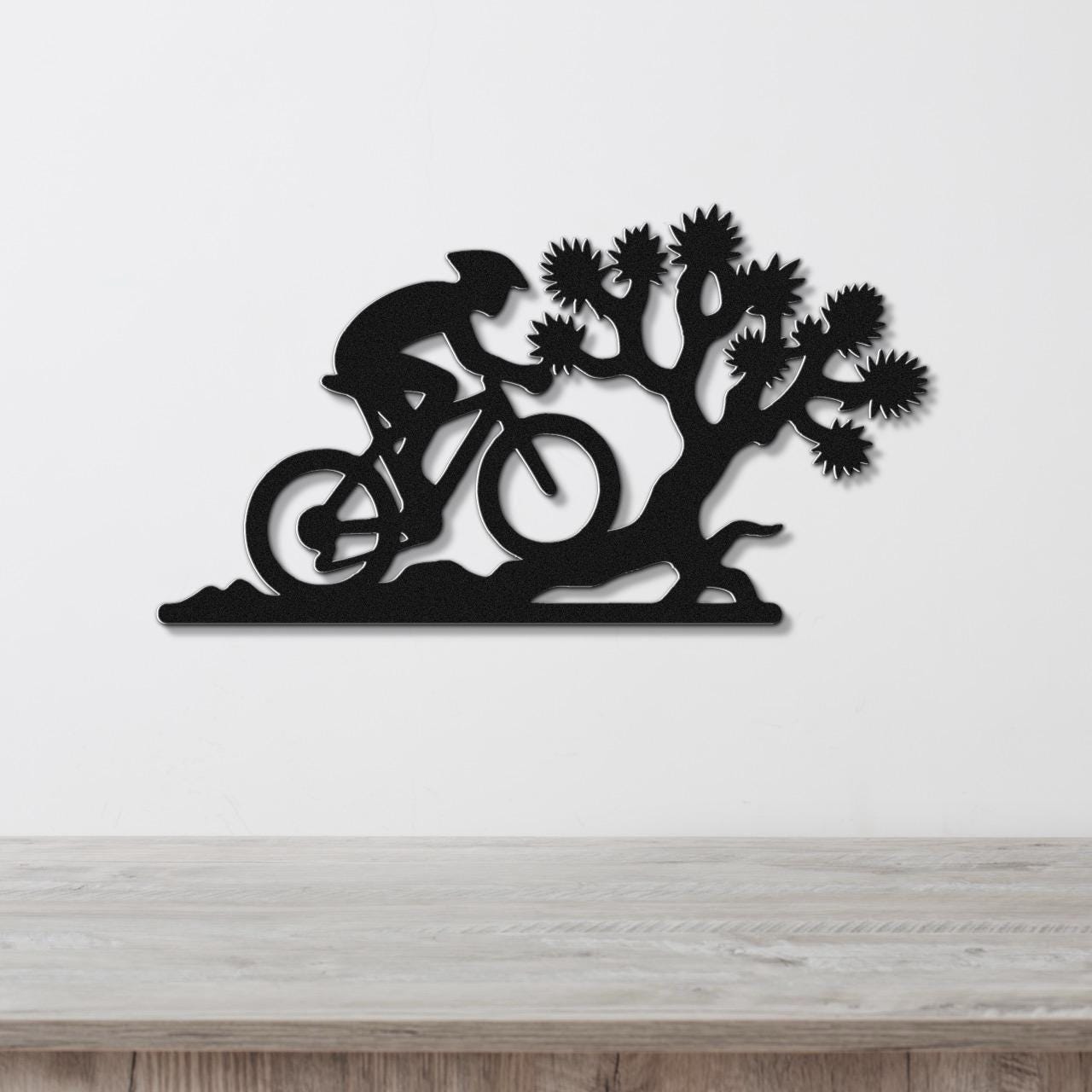 Black metal wall art cutout of a cyclist on a mountain bike beside a Joshua tree, mounted on a white wall above a wooden surface.